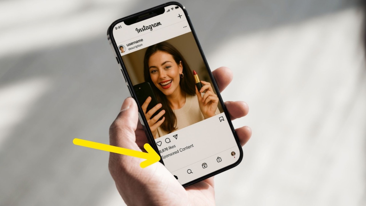Close-up of a hand holding a smartphone displaying an Instagram post of a smiling woman holding red lipstick, with a yellow arrow pointing to the “Sponsored Content” label below the post indicating a paid partnership disclosure.